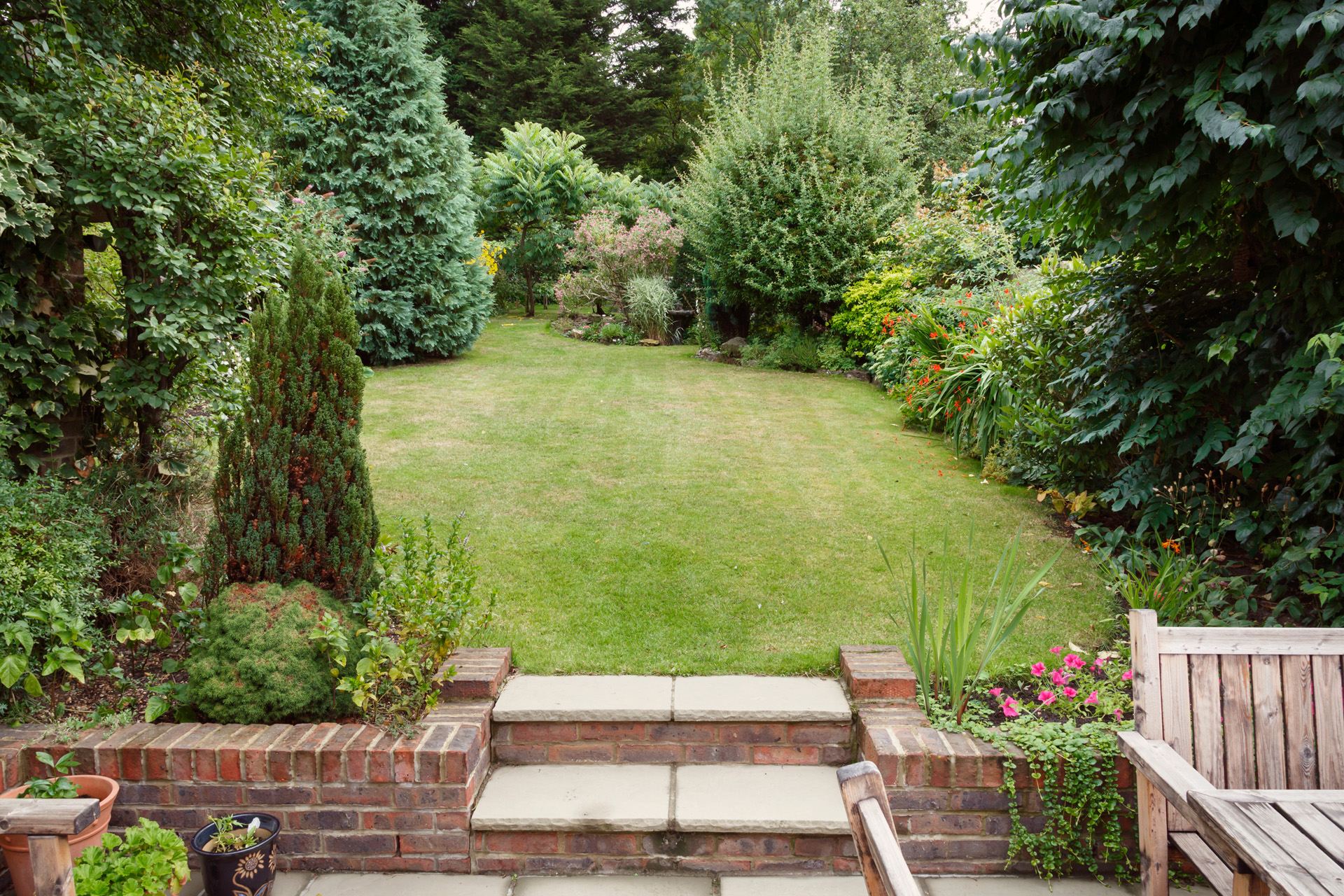 Jardines pequeños con terraza, muebles de jardín y jardineras delante, césped detrás con pequeños árboles y arbustos.