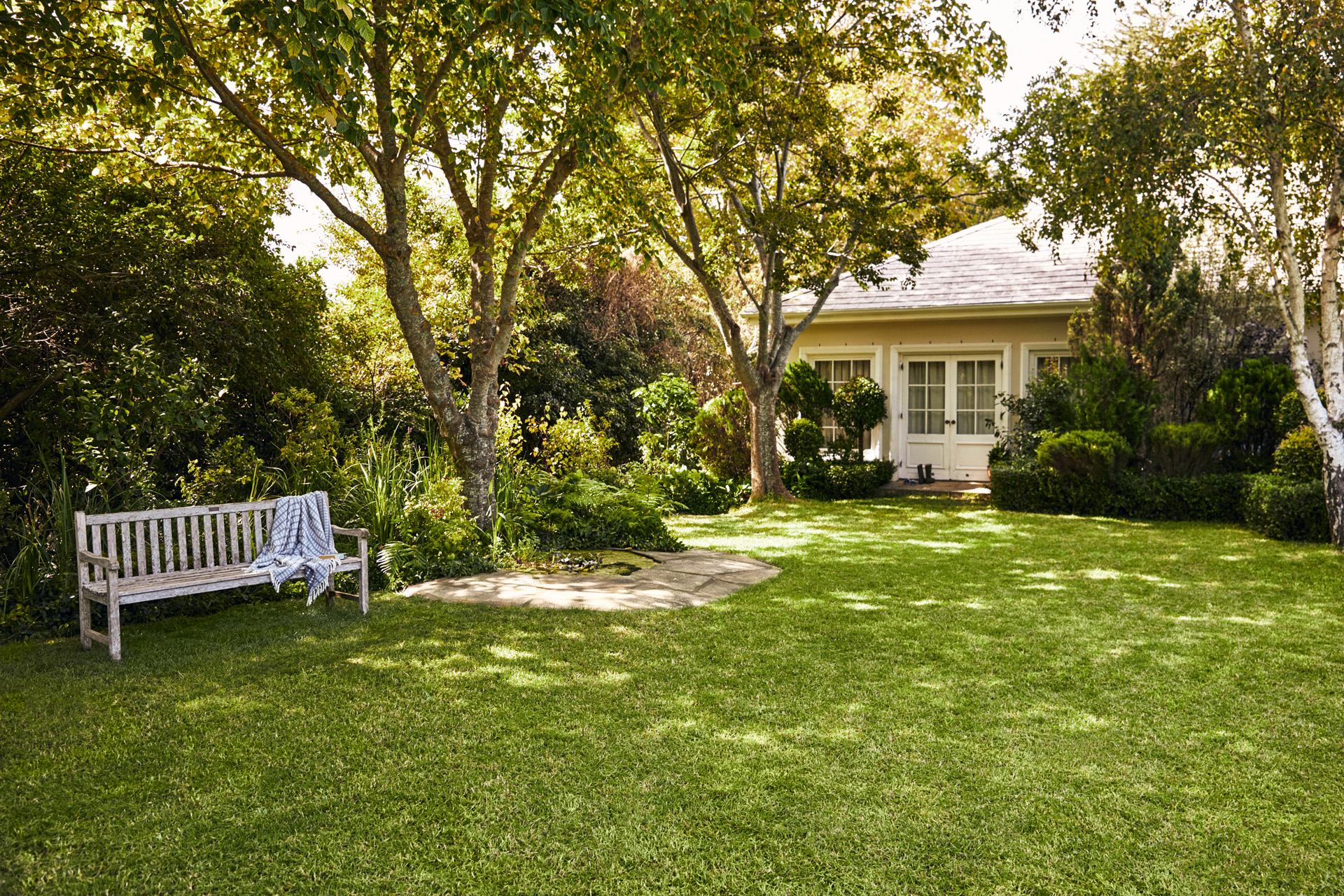 Casa pequeña con jardín, en primer plano un banco de jardín sobre césped verde